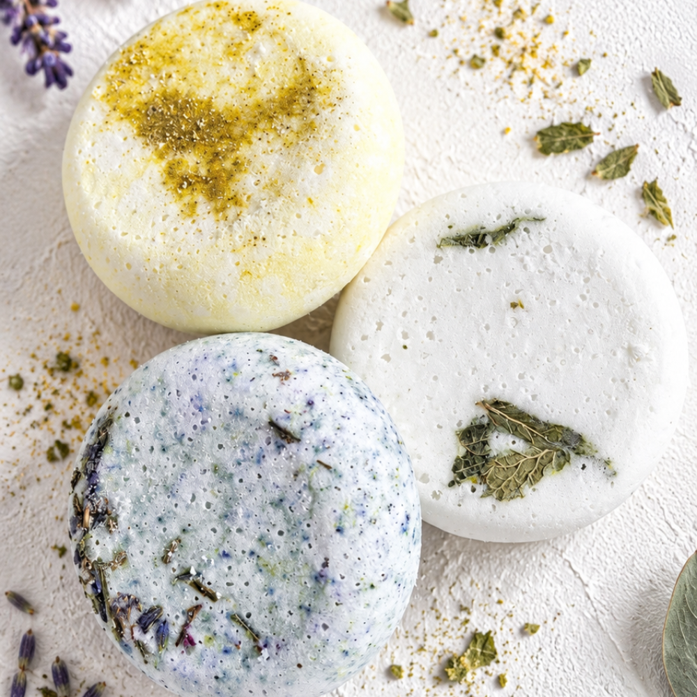 Three round shower steamers with dried botanicals and lavender, arranged on a textured white surface with scattered lavender sprigs and leaves.